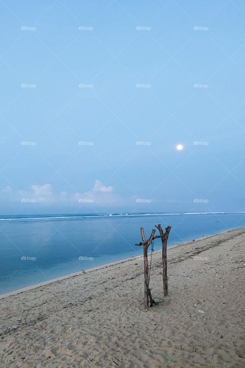 View of a calm beach with clear sea water, blue sky, and the moon visible in the distance