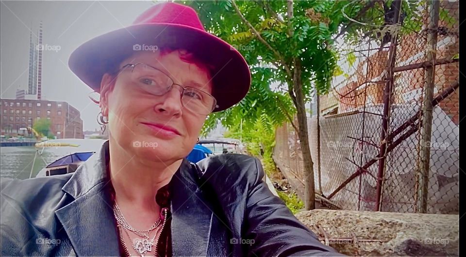 Here I am, “Petra Schiller” smiling to myself standing in the parking lot beneath the “Pulaski Bridge” by the waterfront. The lush green trees are adding to the beauty of this cool Indian summer day. 2022. Hypnotic Productions