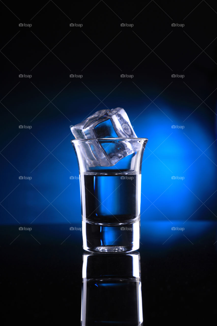 Shot glass with ice cube on blue background