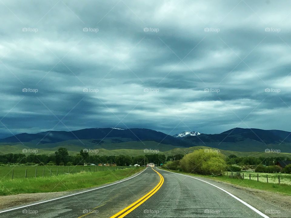 Scenic summer drive on an open country road, featuring: green grass, mature trees, rolling hills, snow-capped mountains, and a grey stormy sky. 