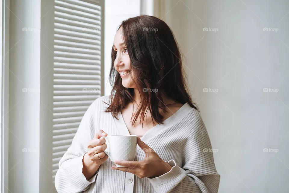 Young beautiful woman forty year with brunette long hair in cozy knitted cardigan with cup of tea in hands in bright interior at home