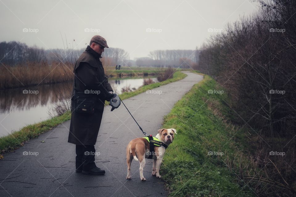 Man cold coat jacket dog bulldog brown white leash water canal walking street way relax exercise enjoy happy stand nature grey sky cloudy recreation outdoor activities 