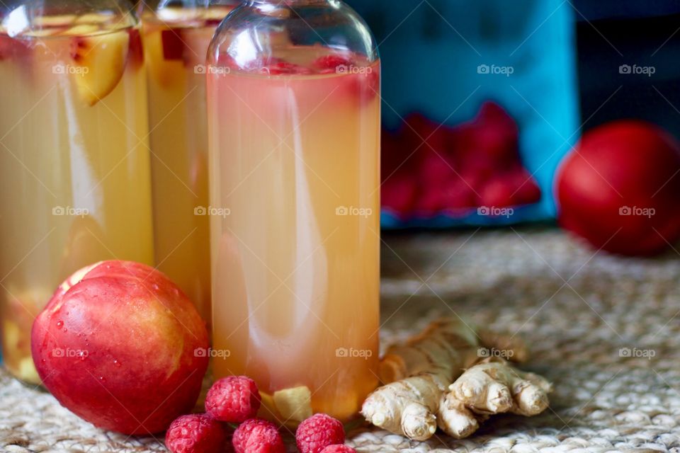 Kombucha, bottled for a second-ferment and flavored with nectarines, raspberries and slices of ginger root on a natural fiber mat, whole nectarines and raspberries in a paper carton in the background