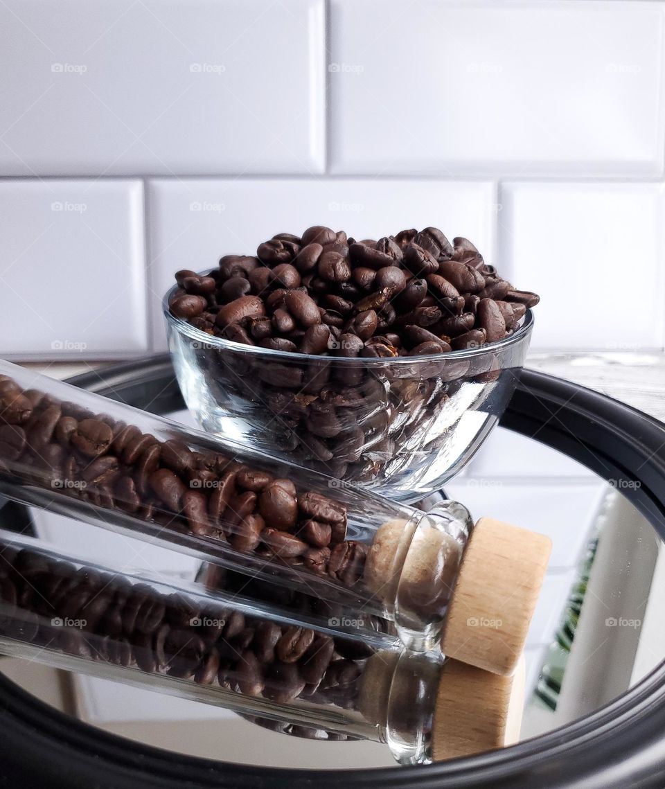 A glass bowl of coffee beans and a coffee beans glass container on a mirror tray with a white tile kitchen background.