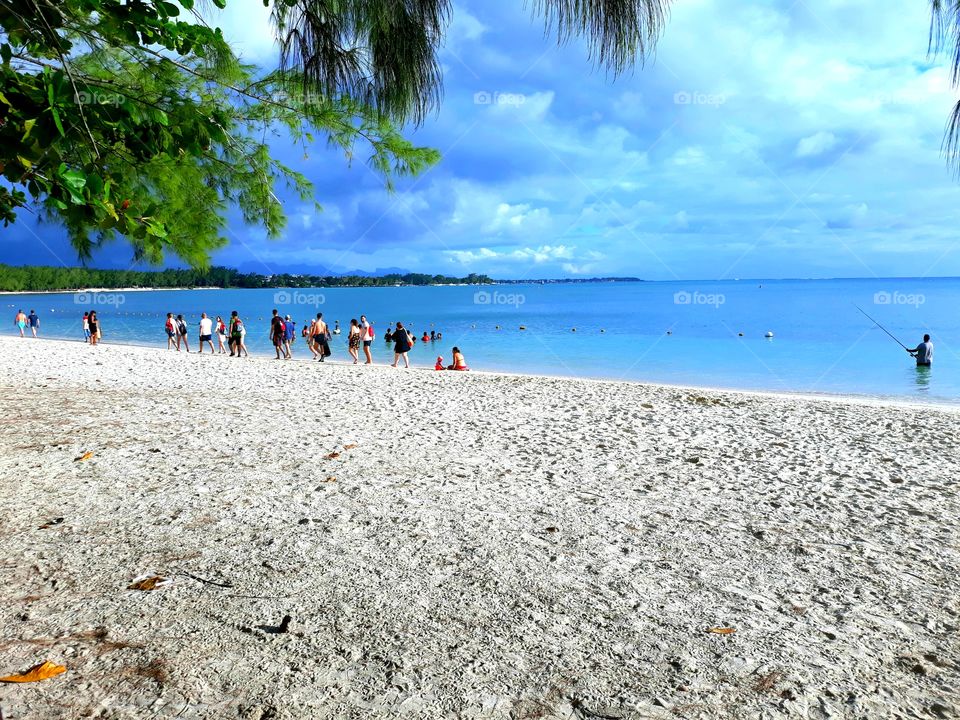 Beach with nice white sand everywhere, blue Sea, beautiful Sky, tourists walking across the seashore, fisherman fishing with the body inside the water and green leaves.