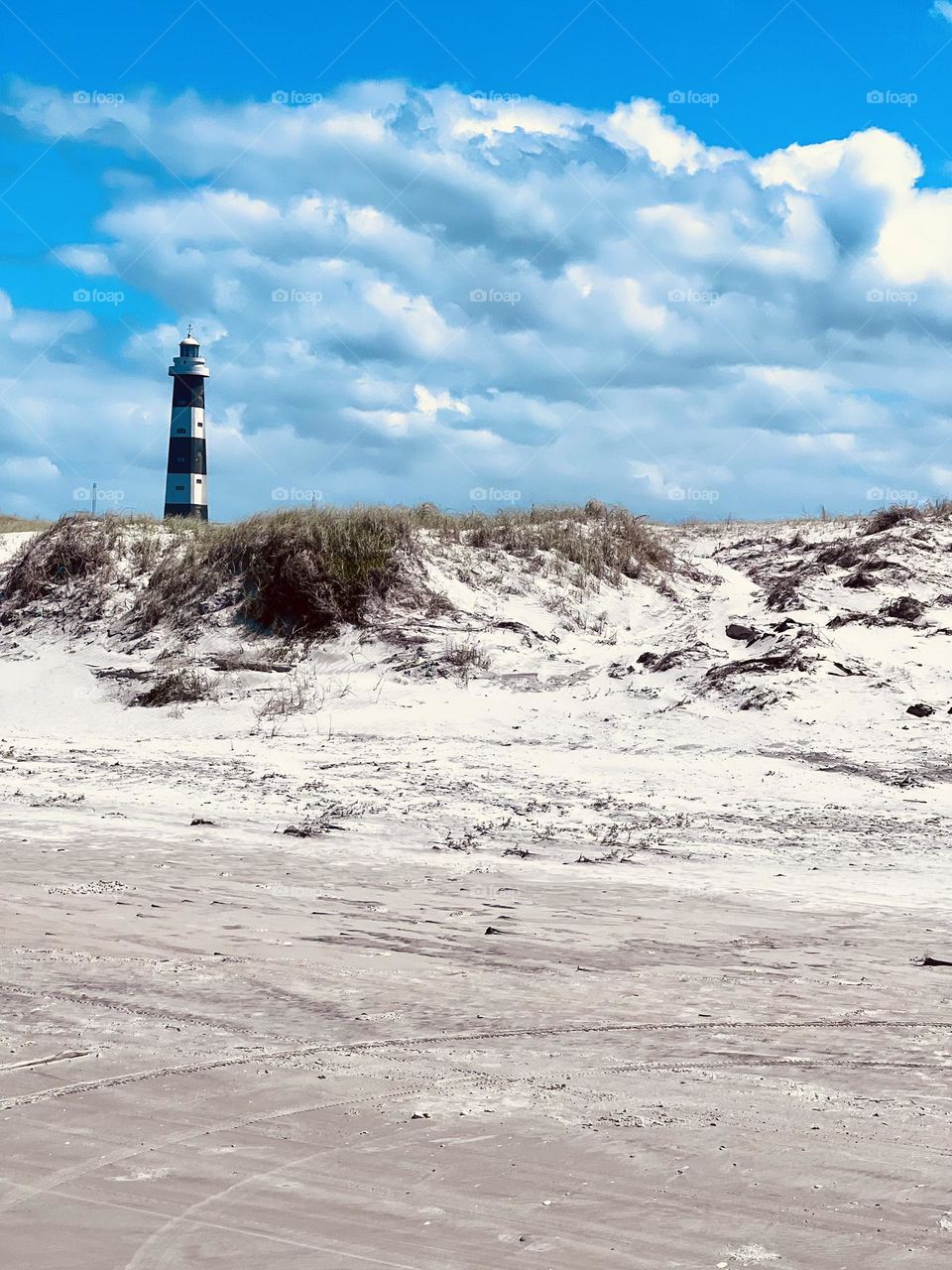 A blue and white striped lighthouse stands majestically on a light sand beach, surrounded by grasses and under a blue sky with white clouds, evoking a feeling of tranquility and serenity.
