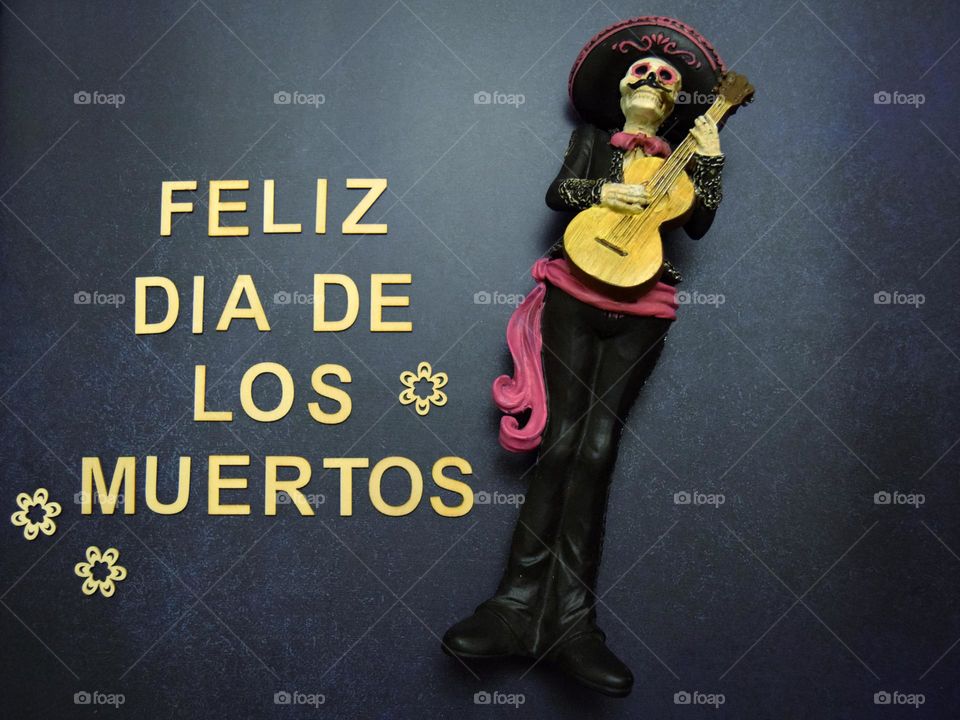 A simple phrase flatlay which reads “Feliz Dia De Los Muertos” accompanied by a mariachi man wearing pink & black traditional dress, with the addition of decorative wooden flowers.