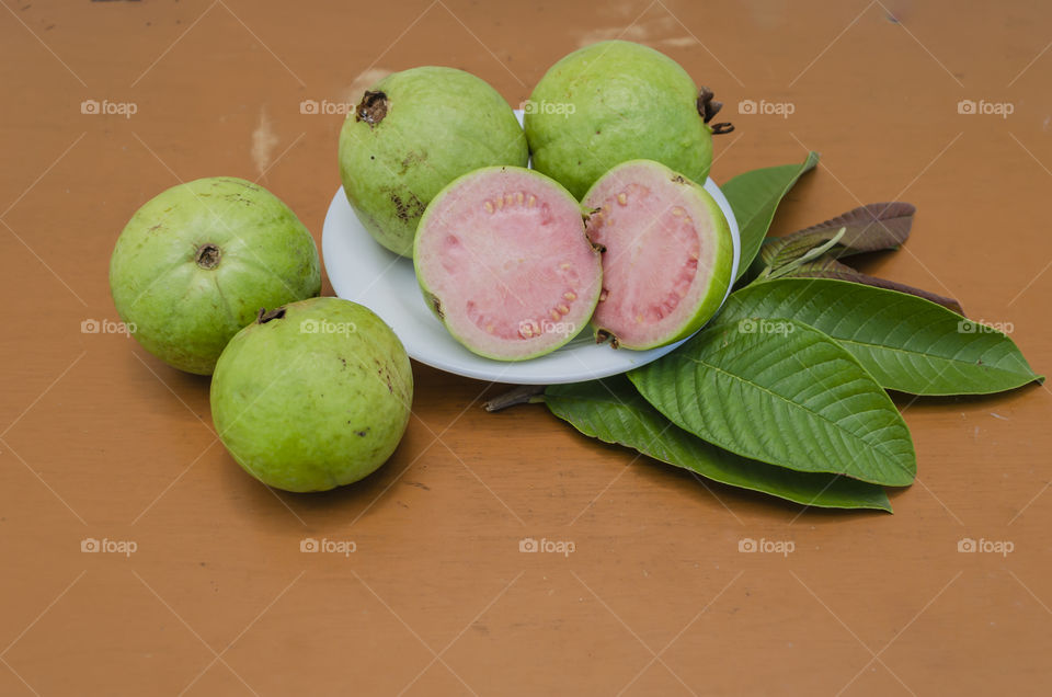 Cut and whole guavas in a plate on tope of leaves, that are resting on a brown table top with other of the fruits beside the white plate.