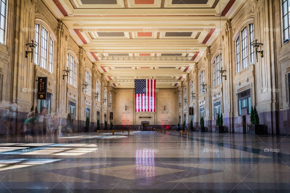 Horizontal photo of the interior of Union Station in Kansas City, MO with a large American flag on the back wall and people blurred out through a long exposure
