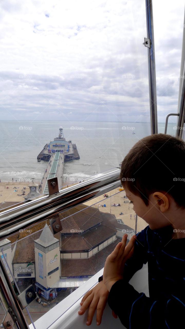 Boy on ferris wheel looking down and waving happily view of sea and pier at background 