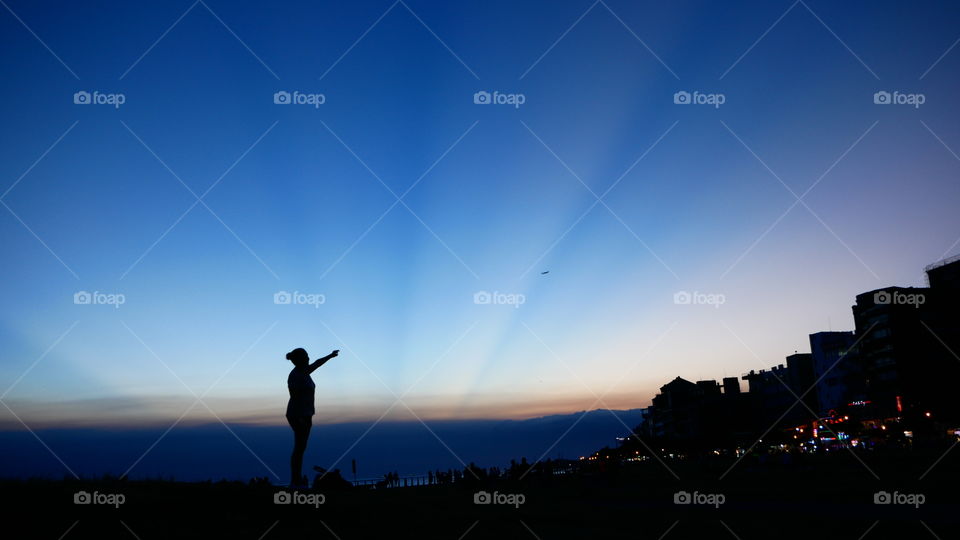 Reaching for the sky. A woman silhouette reaching for the sky. A plane flying in the Distance. Sunray at dusk are seen in the background. Dark blue calming colors.