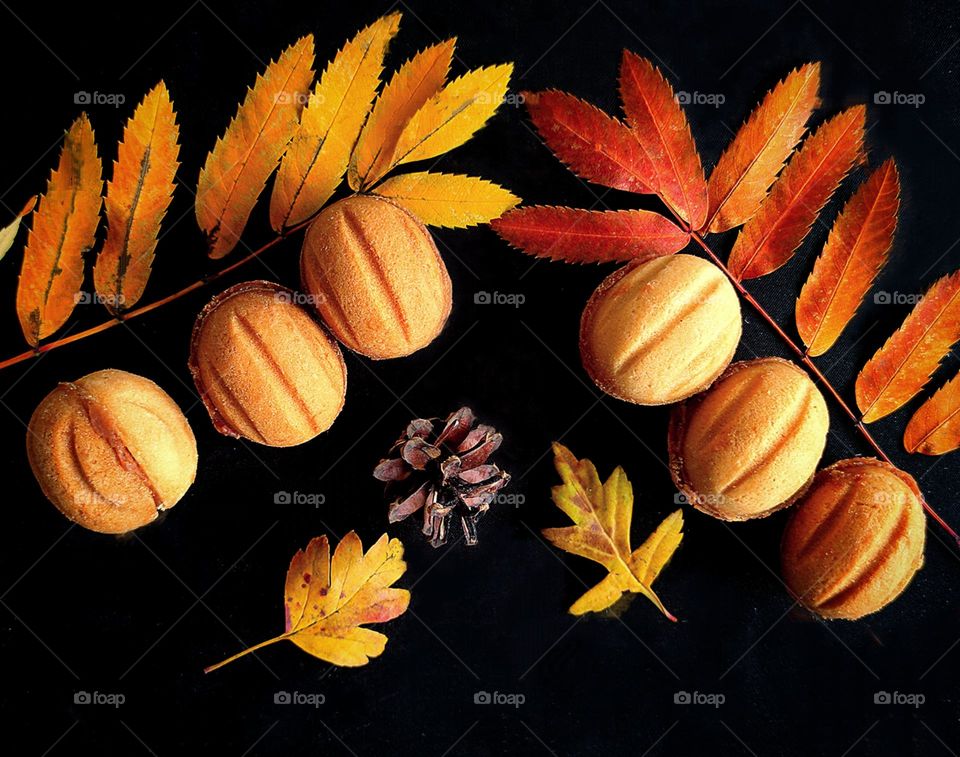 On a black background are: a branch of a tree with red leaves, under which there are three cookies in the form of nuts; a branch of a tree with yellow leaves, under which lie three cookies in the form of nuts; yellow leaves and a pine cone