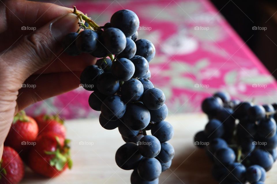 A man sitting at a kitchen table holds a stem  filled batch of sweet grapes in his hand.