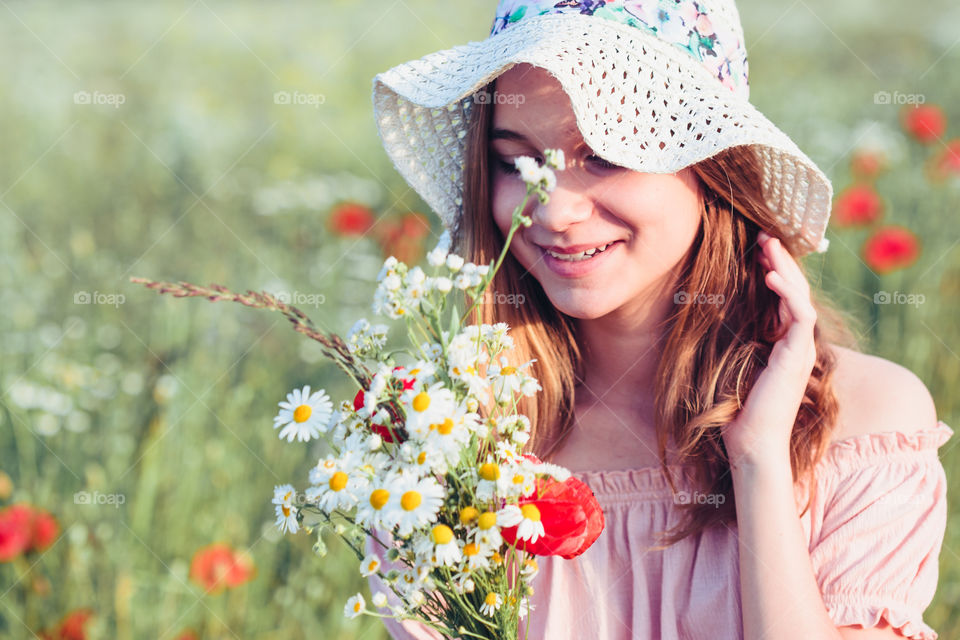 Beautieful young girl in the field of wild flowers. Teenage girl picking the spring flowers in the meadow, holding bouquet of flowers. She wearing hat and summer clothes. Spending time close to nature