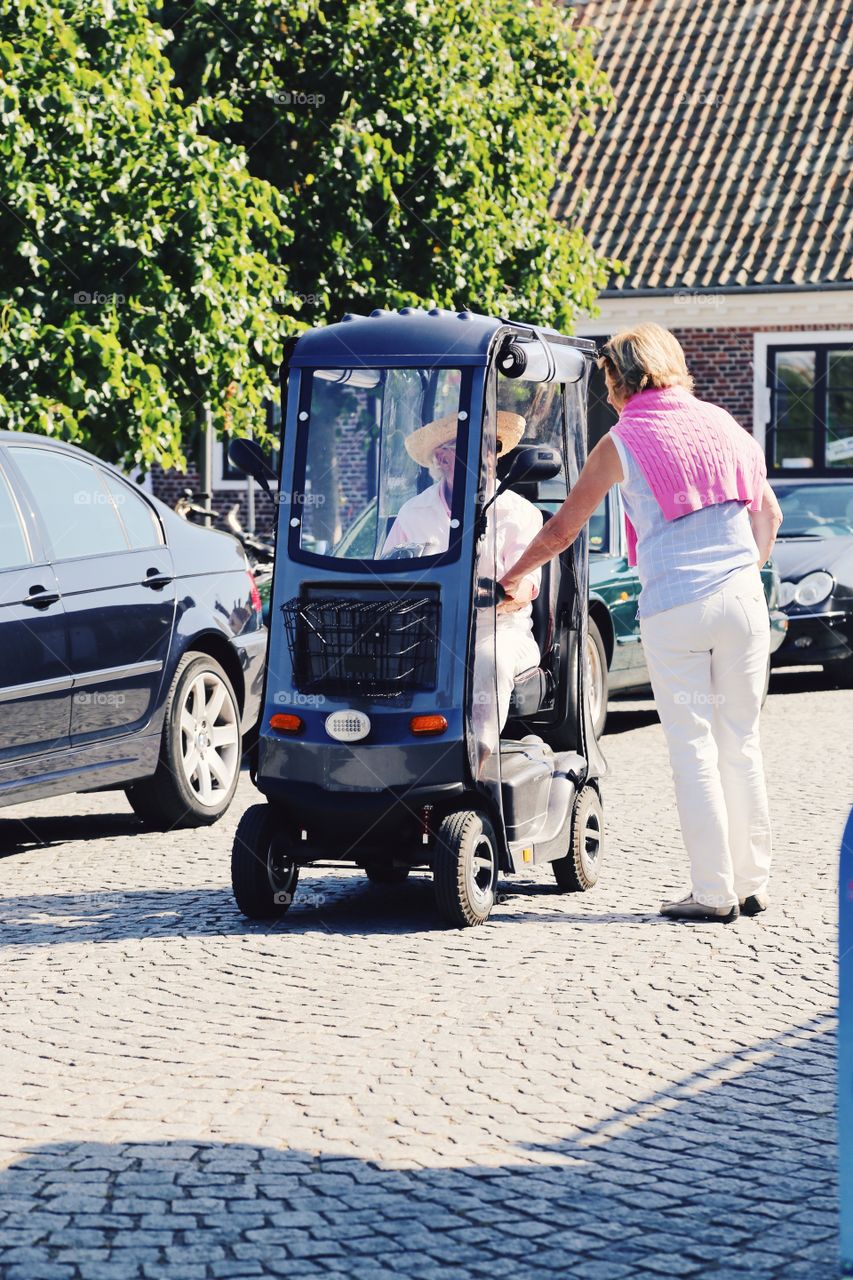 Two people standing on the small pebbled stone street. One one is sitting in a covered vehicle on four small wheels. Cars are parked on the side.