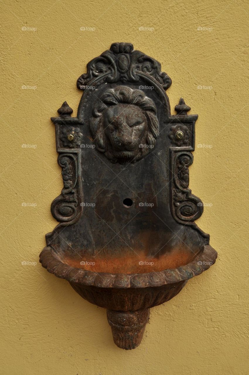 Metal garden rusted fountain decorated with lion head against yellow painted wall 