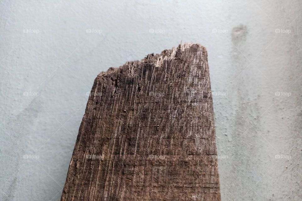 Close-up of a worn-looking, light brown piece of wood against a white wall background. The wood has a rough texture and clearly visible wood grain