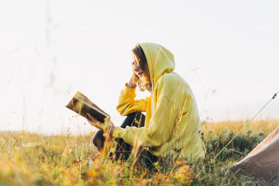 Young woman traveler in yellow hoodie reading book against tent. Local tourism, weekend trip