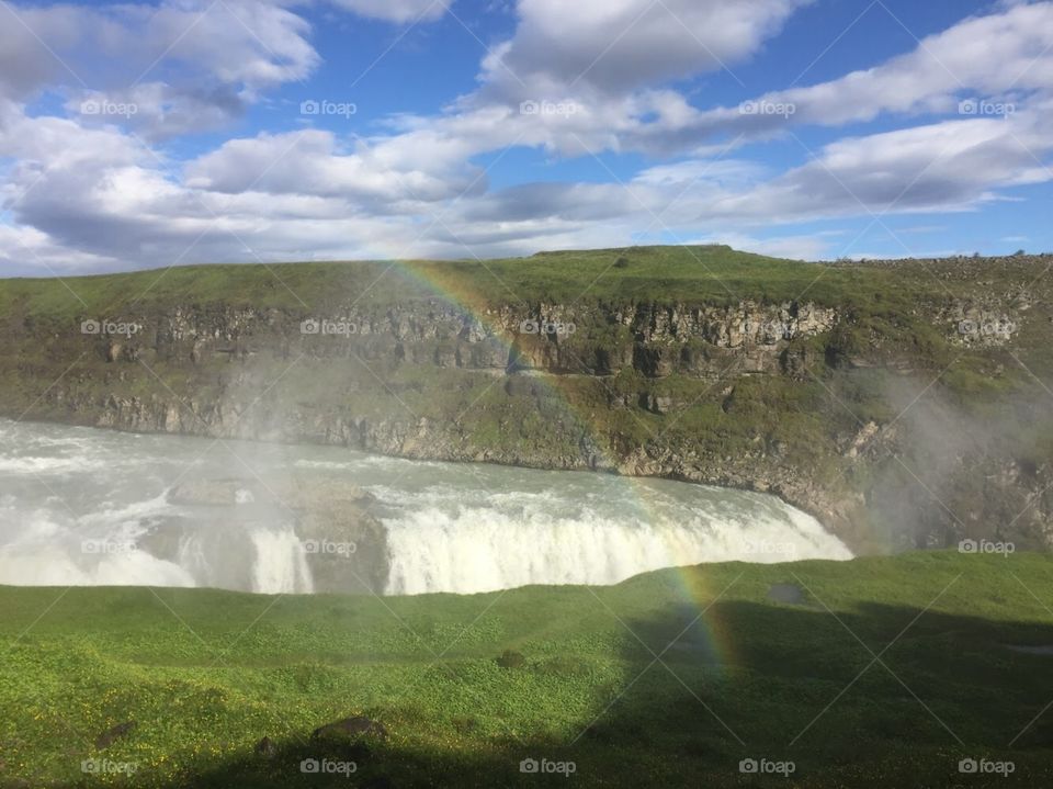Golden Circle Waterfall in Iceland. Large amounts of rushing water with rising steam, clouds, a blue sky, and green mountains in the background. Large rainbow! 