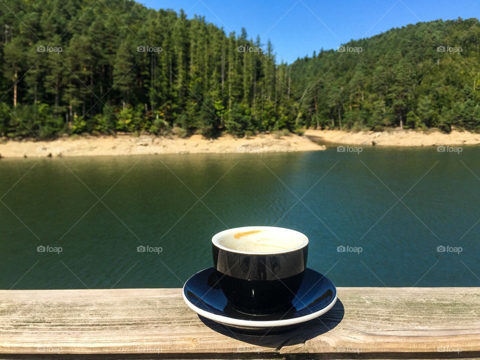 Cup of coffe on plate served on a terrace near a lake surrounded by forest