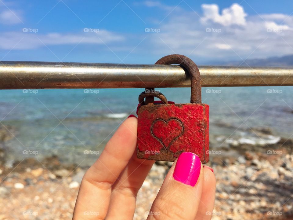 Woman hand wearing pink nail polish holding a red padlock on a bridge over the sea