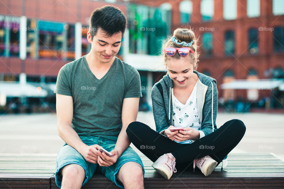 Couple of friends, teenage girl and boy,  having fun together, using smartphones,  sitting in center of town, spending time together. Real people, authentic situations