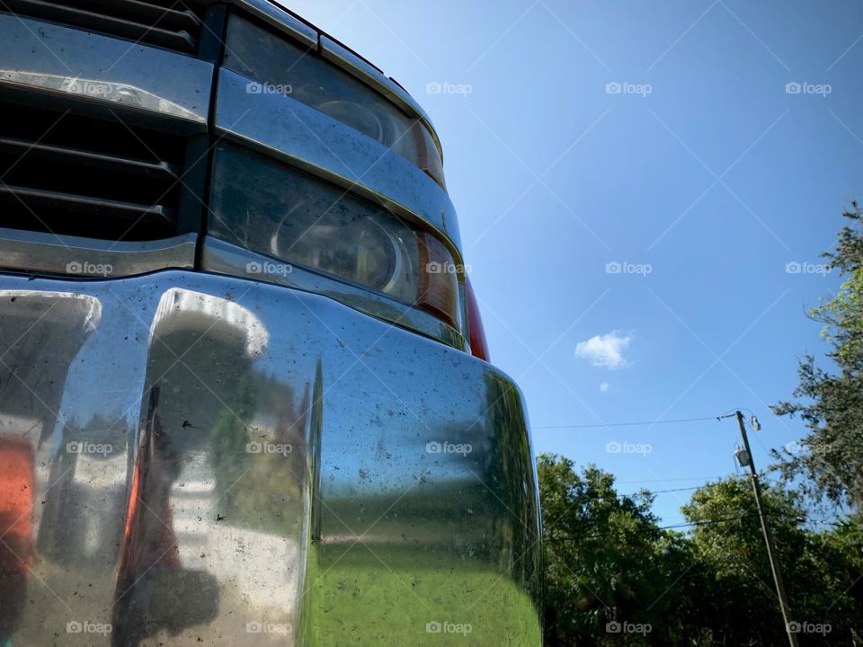 Commuting: front left side of a red pickup truck vehicle with chromed front bumper reflecting as a mirror the green trees, grass and home entrance, fence, close up.