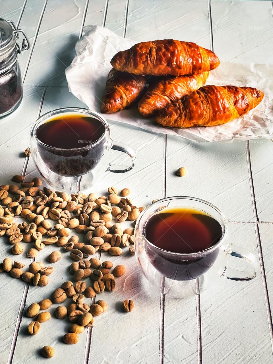 Hot black coffee in cups, some scattered coffee beans and croissants 