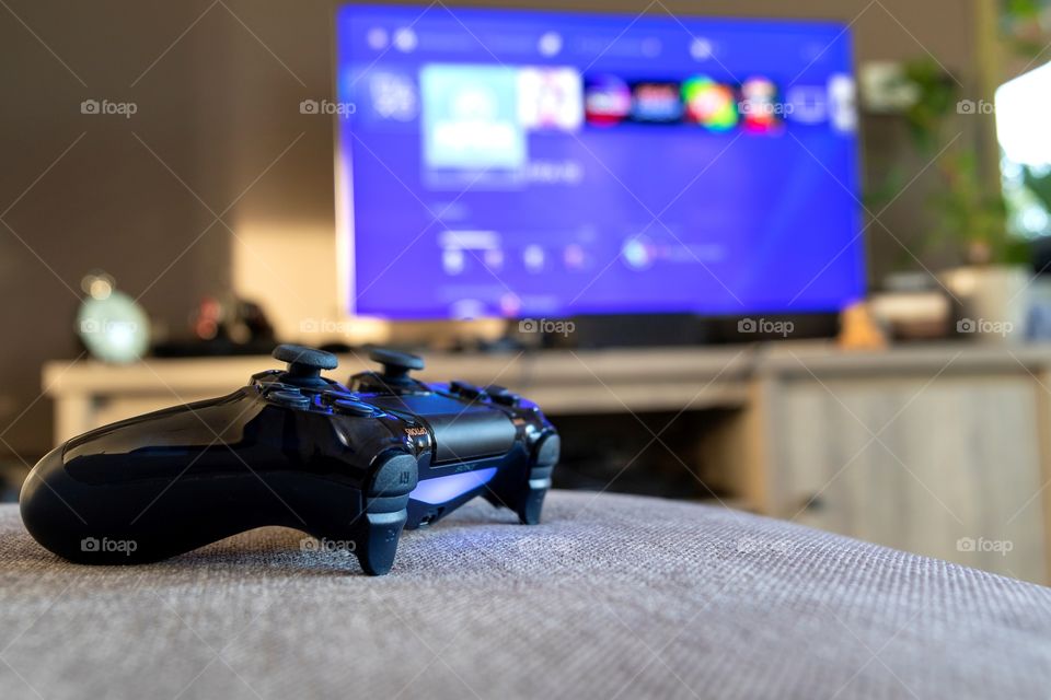 A portrait of a playstation 4 controller in front of a tv with the playstation home screen blurred in the background.