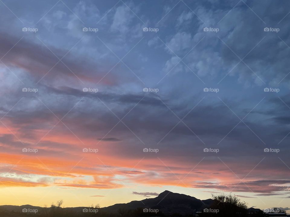 A yellow, pink, and blue sunset with mountains and tree branches below.