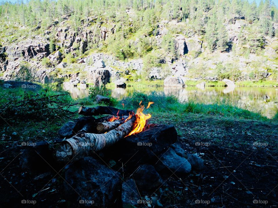 Campfire with birch logs and the morning sun to the river on the otherside
