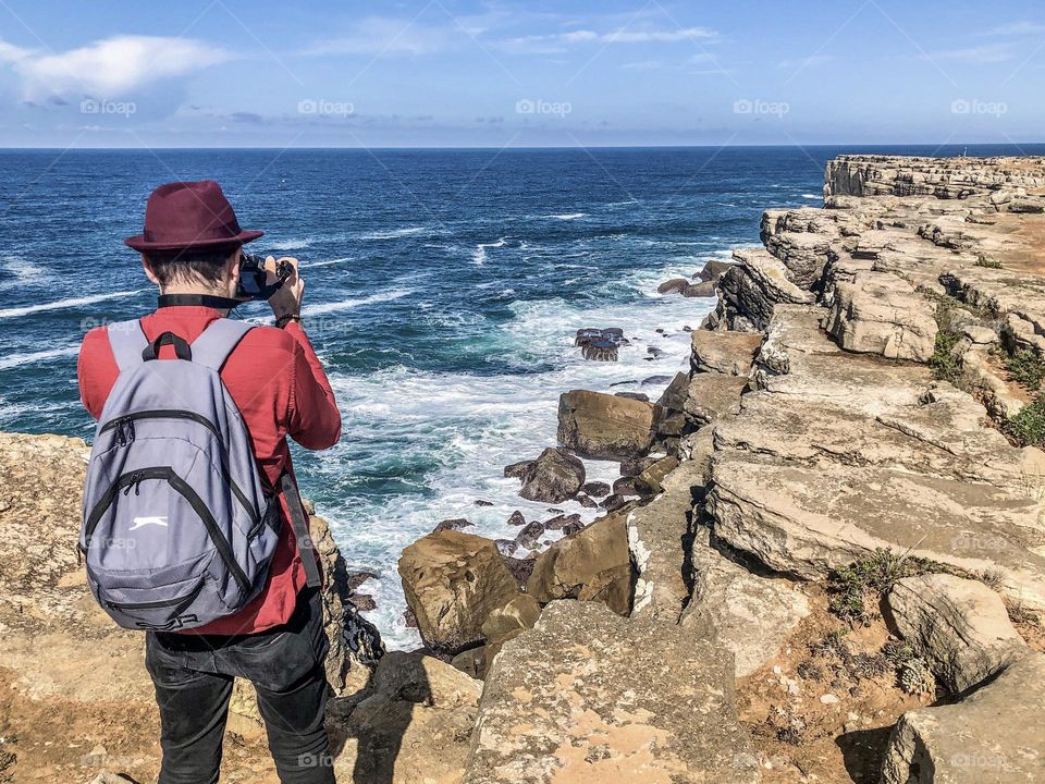 A backpacker stands on cliffs taking pictures of the rocky coastline at Peniche, Portugal 