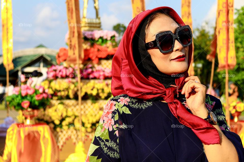 Portrait of a beautiful mother wearing a red hijab, wearing a floral patterned robe and wearing black glasses, taking a photo with a happy expression.