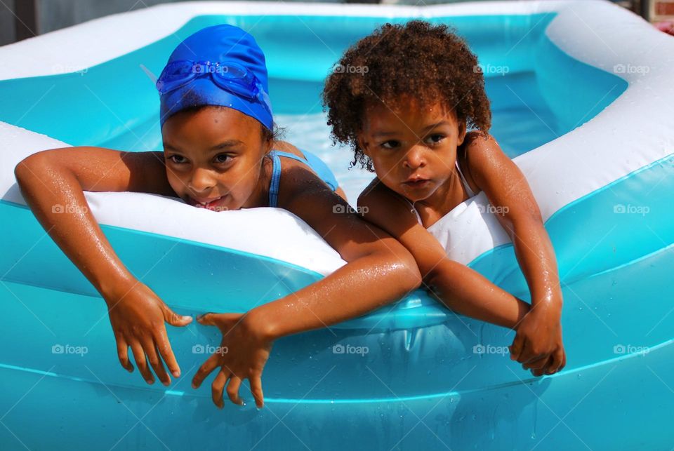 Girl of mixed race enjoying the refreshment of water in a swimming pool on a hot summer day, together with her little sister (family, fun, summer, water, blue, swimming suit, splash, hot, enjoy, play, outdoors)