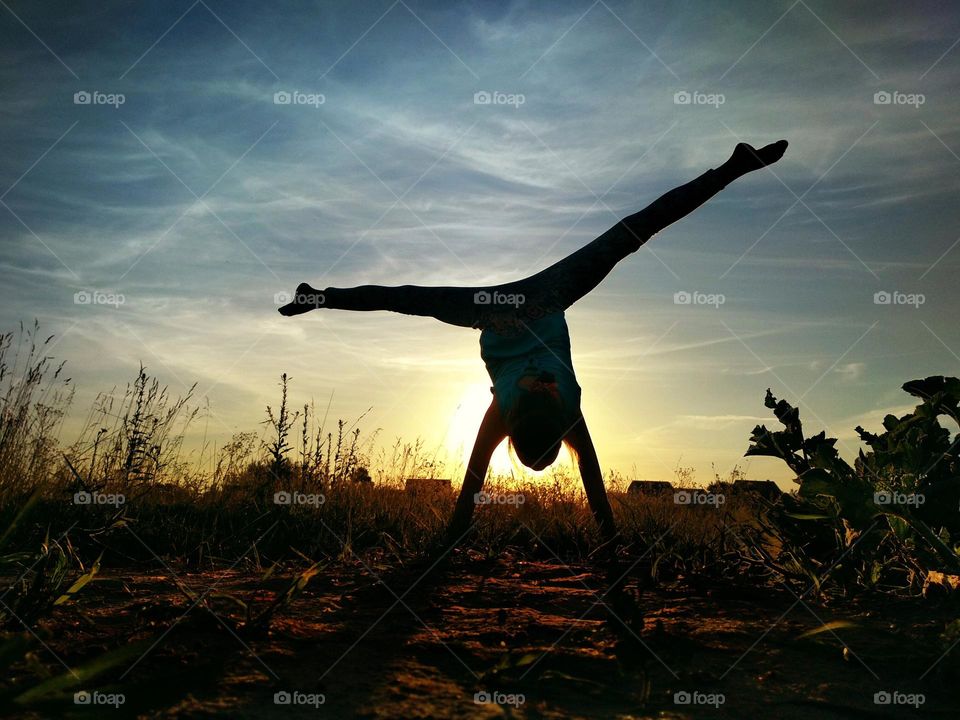 Girl doing split on handstand. A model stands on her hands, doing gymnastic splits against the blue sky. Healthy lifestyle concept. Silhouette on sunset background. Gymnastic in nature