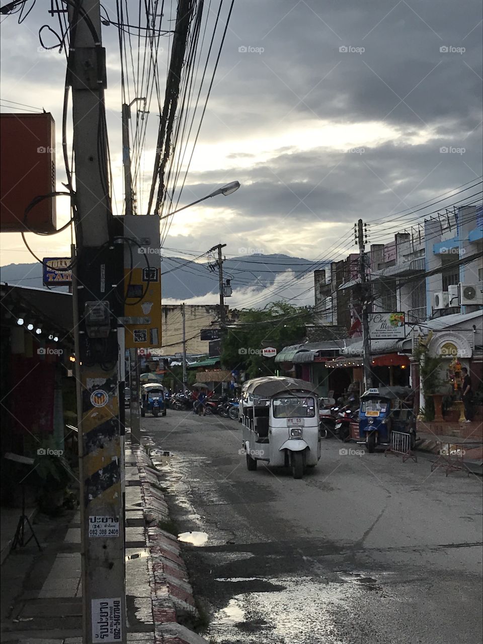 Late afternoon, Chiang Mai, Thailand. The sun sets over the mountains. Storms are coming. A tuk-tuk droves down a street. The famous Asian telephone lines cover the sky. 