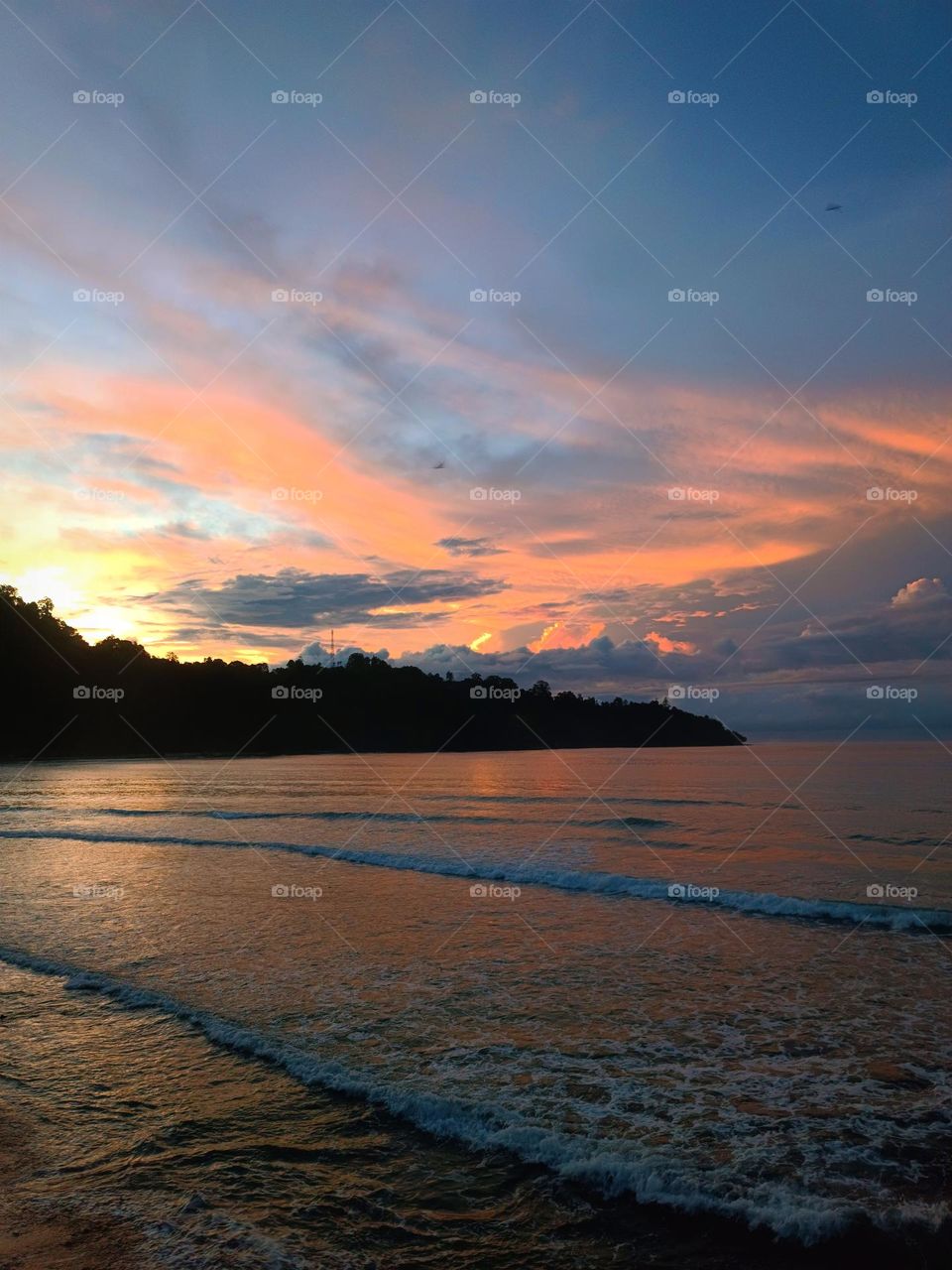 beautiful clouds in morning with waves breaking in foreground and silhouette of mountain
