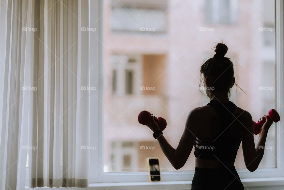 Young woman working out at home watching a workout lifting weights near a window