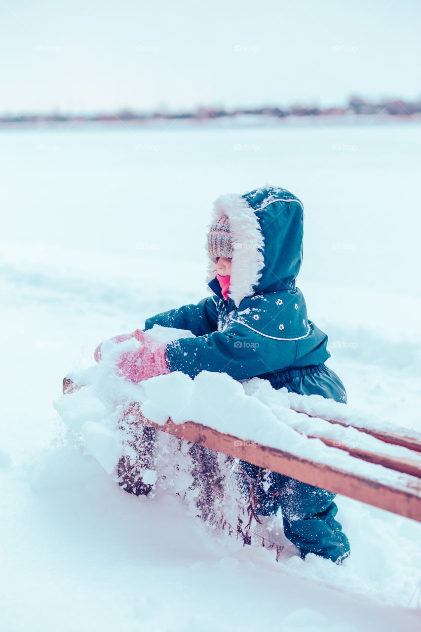 Little girl enjoying winter removing snow from a bench. Toddler is playing outdoors while after snowfall. Child is wearing dark blue snowsuit and wool cap