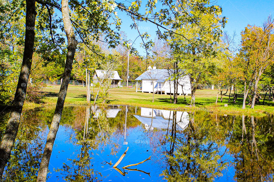 Two historical buildings in a park reflected in a clear blue lake alomg with green trees under an autumn sky..