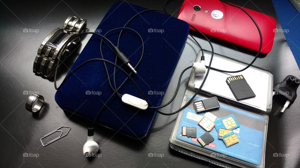 Mesa de escritório, com fone de ouvido, cartões de memória, chips de celulares, carteira, cartões de créditos, anel, relógio, clipe, smartphone...