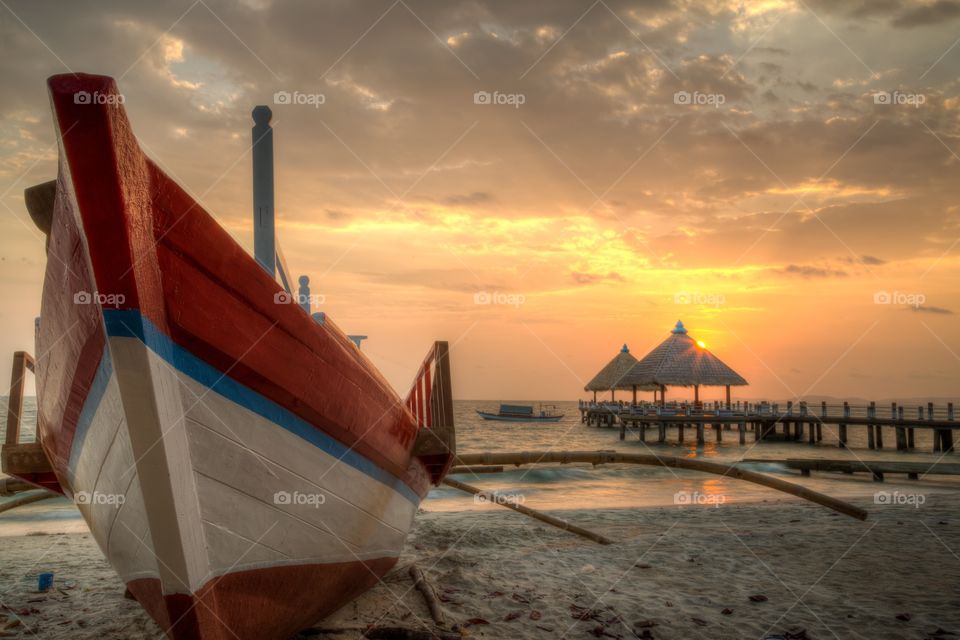 Cambodian Orange beach sunset . Orange sunset at the beach next to the sea. Fishing boat on the shore. Couple straw huts on pier. One fishing boat