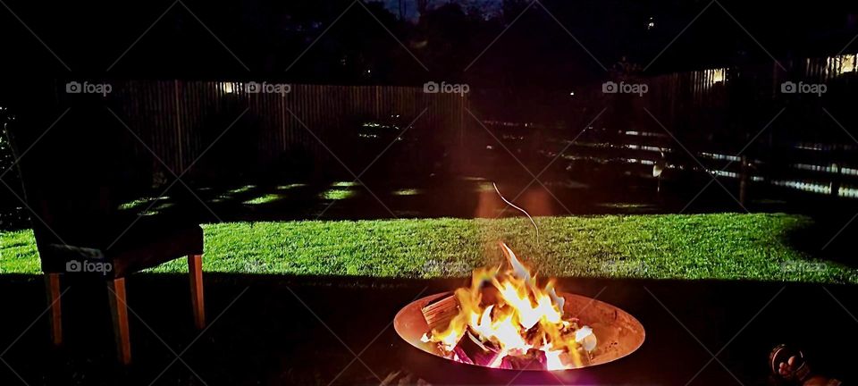 Another view of the bonfire at night and the garden in which it is located in “Bavaria”, Germany. A spotlight shows the green lawn clearly at this late hour when it is dark outside. 2023. Hypnotic Productions