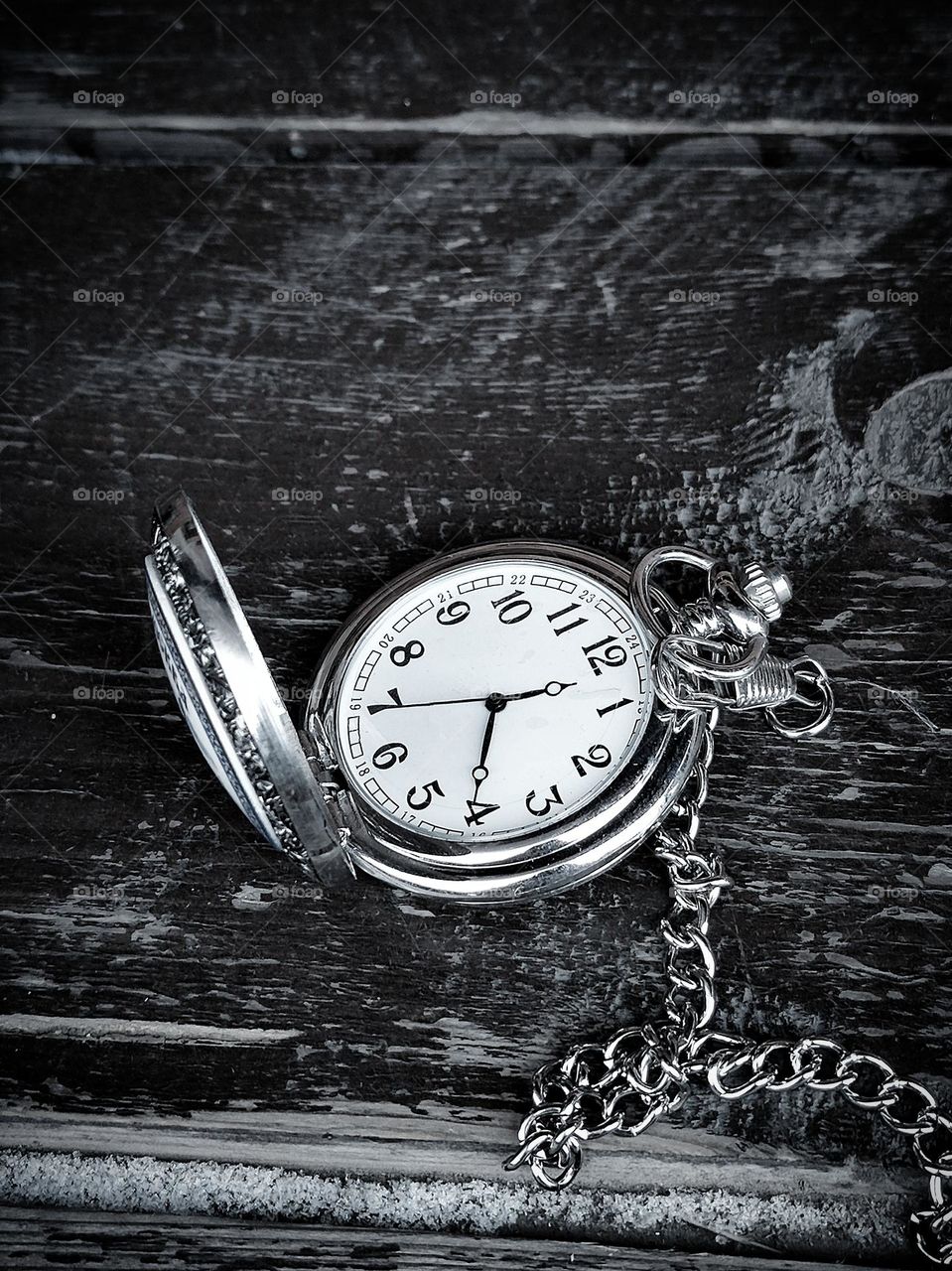 Black and white photo.  On a wooden background lies a vintage style pocket watch with an open lid and a metal chain