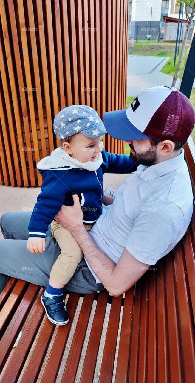 The father is sitting on the bench, hugging his baby, talking lovingly and making him happy.