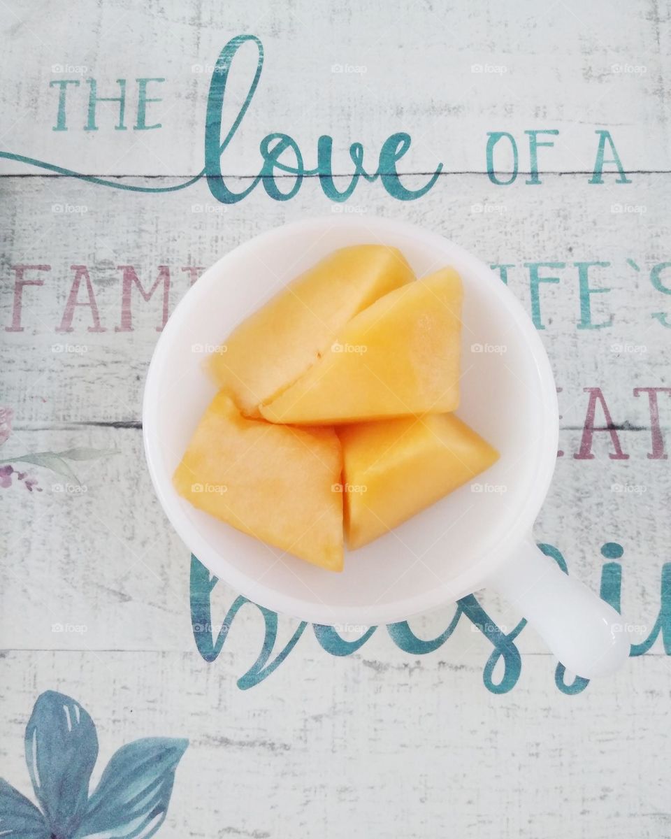 A white coffee mug with pieces of cantaloupe in it on top of a place mat that says " The love of a family is life's greatest blessing"