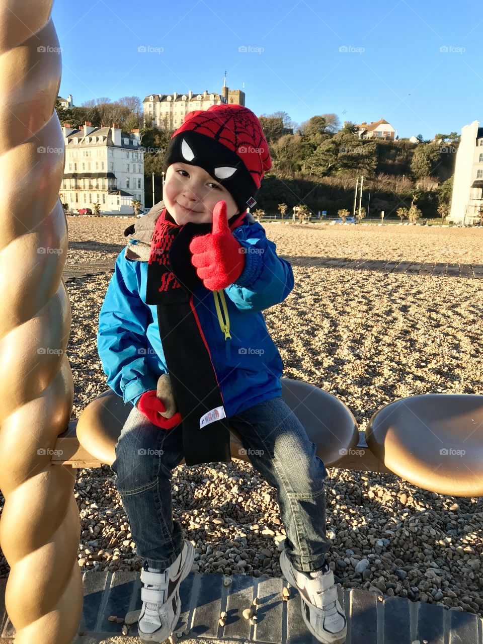 Happy little boy giving a thumbs up on a pebbled beach on a brisk winter’s day