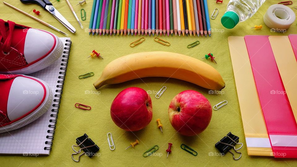 School time! School supplies with a snack and a bottle of water on the background of green paper. Sneakers on a spiral copybook, a banana, apples, buttons, colored pencils, red and yellow folders, paper clips, tape, a bottle of water, paint brushes
