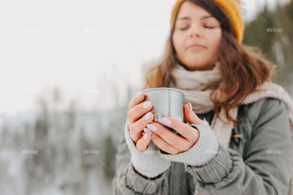 Brunette girl in yellow knitted hat with metal mug of hot tea in the forest outdoors in winter