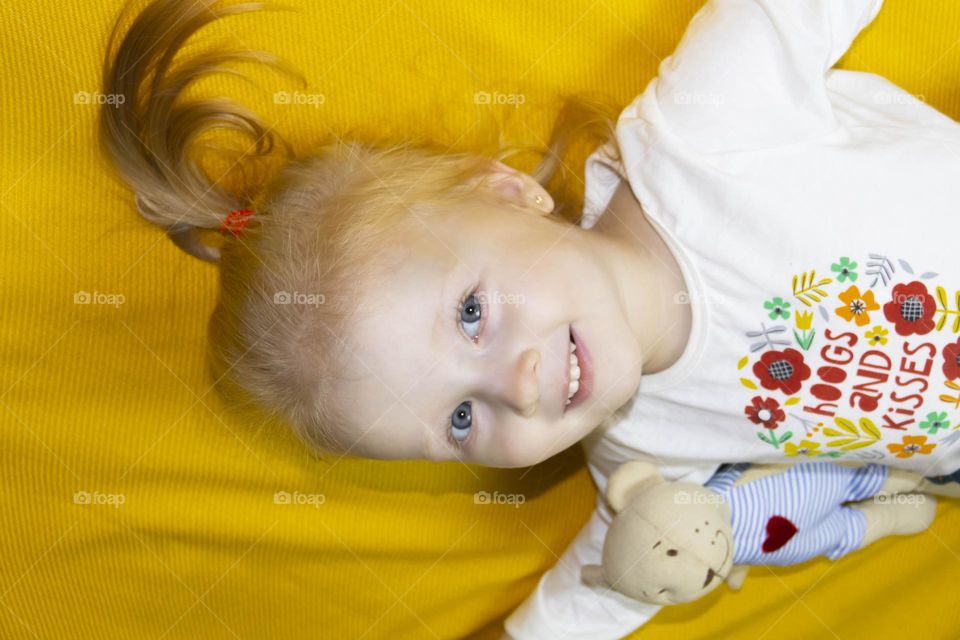 A small, red-haired girl is laughing at home on a gray sofa with purple pillows and a yellow background